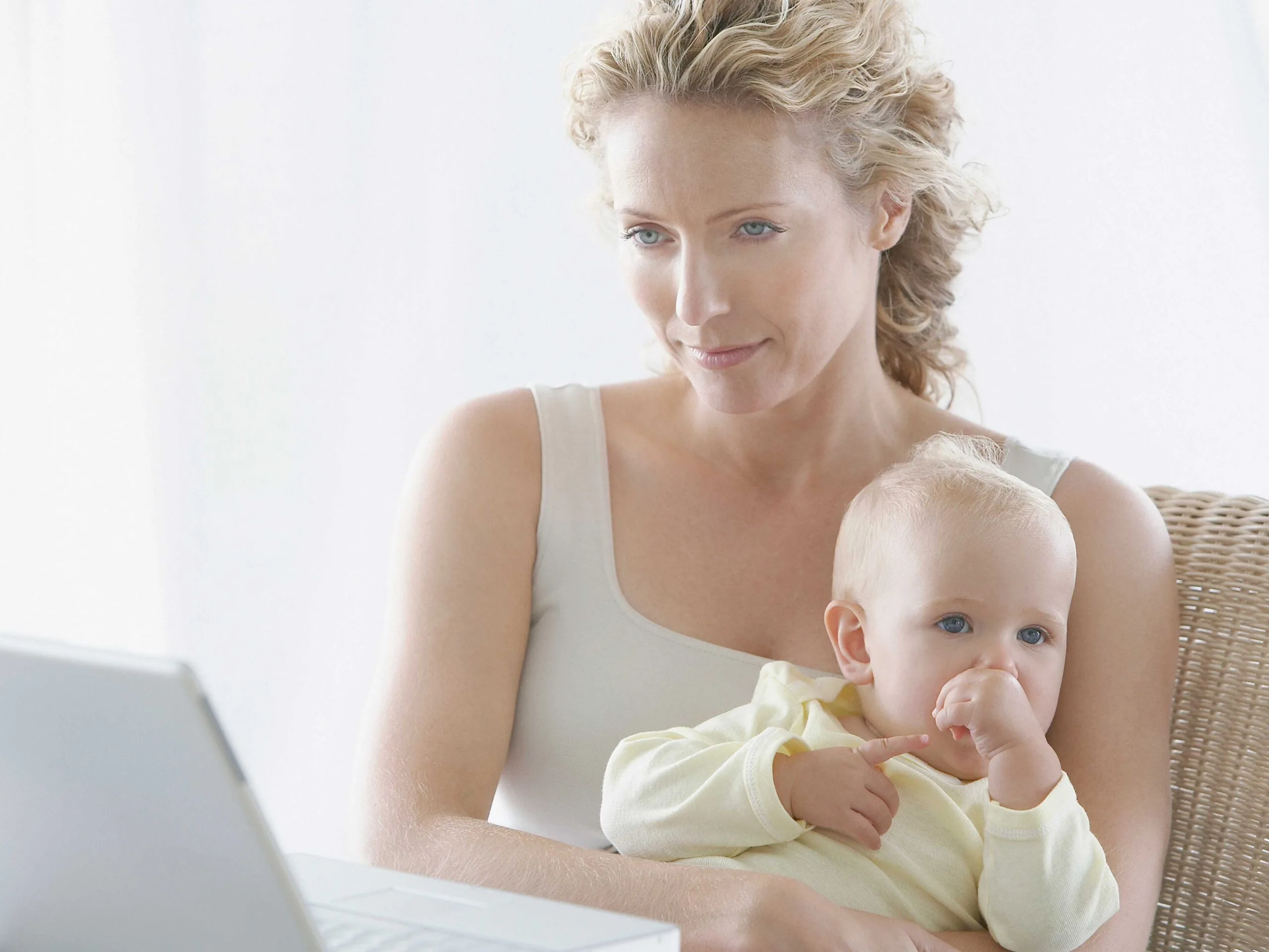 Mother working on a laptop while holding her baby, who is sucking their thumb.