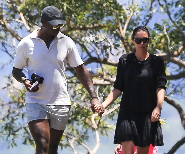 A man in a white shirt and cap holds hands with a woman in a black dress, walking outdoors on a sunny day.