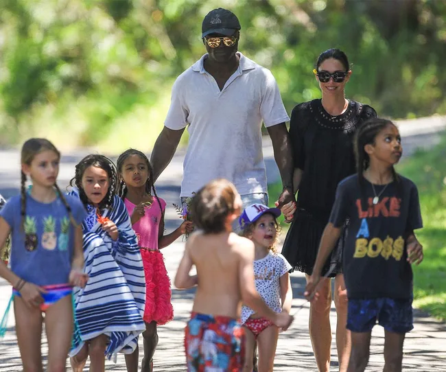 A group of children walking on a sunny day, accompanied by two adults wearing casual clothing and sunglasses.