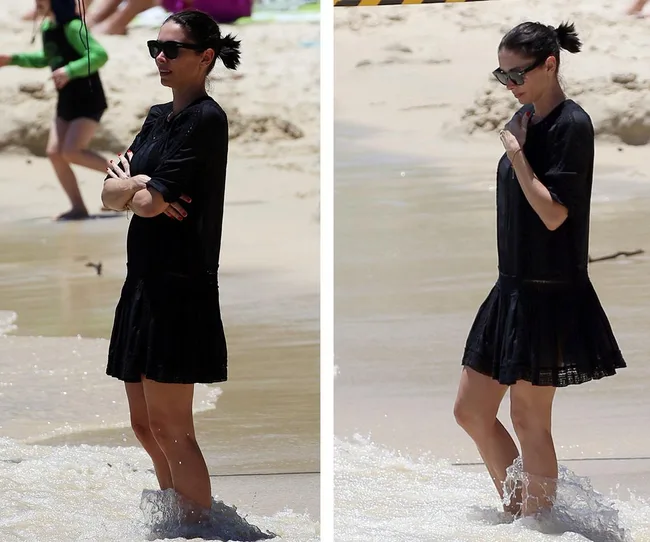 Woman in black dress and sunglasses standing at the beach, water covering her feet, with people in the background.