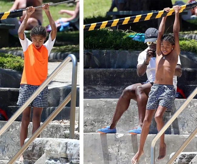 A child in checkered shorts uses a horizontal bar outdoors while an adult watches from nearby steps.