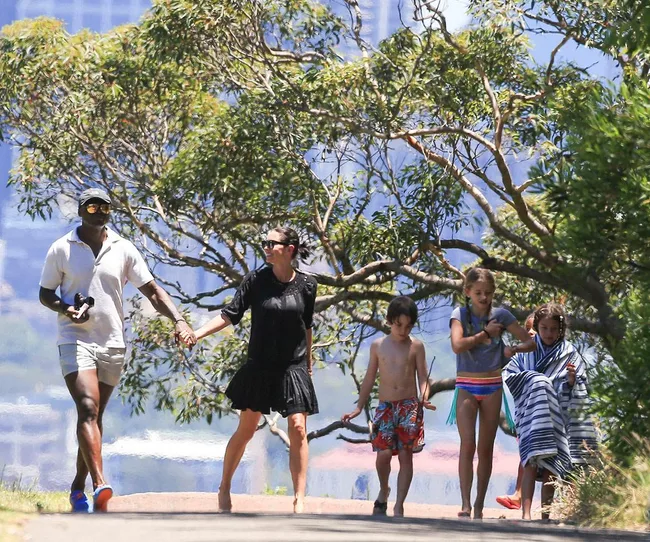 A couple walks on a path with three children on a sunny day, surrounded by trees, two children are wrapped in towels.