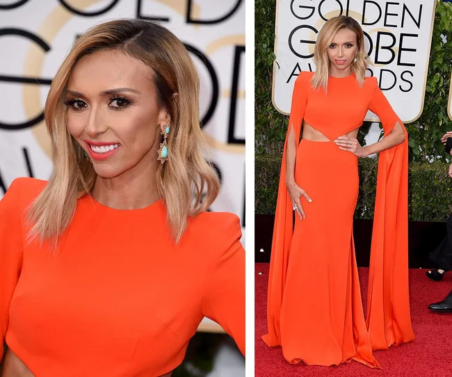 Red carpet event; woman in vibrant orange gown with cape sleeves, turquoise earrings, smiling and posing confidently.