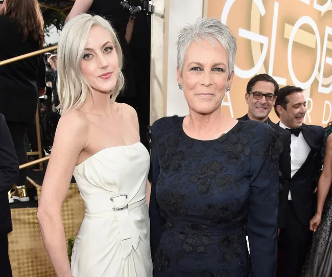 Two women pose at a formal event, one in a white dress and the other in a black floral outfit, with people in the background.