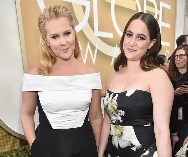 Two women pose together at a formal event, wearing elegant off-the-shoulder dresses with floral and monochrome designs.