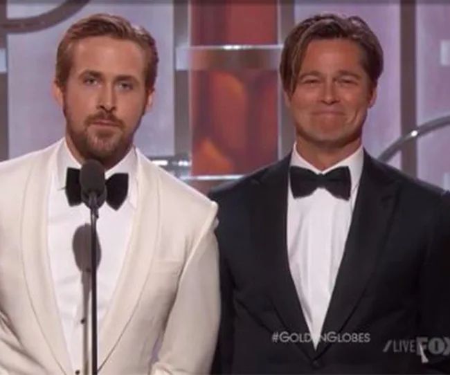 Two men in tuxedos stand on stage, one in white and one in black, at the Golden Globes event.