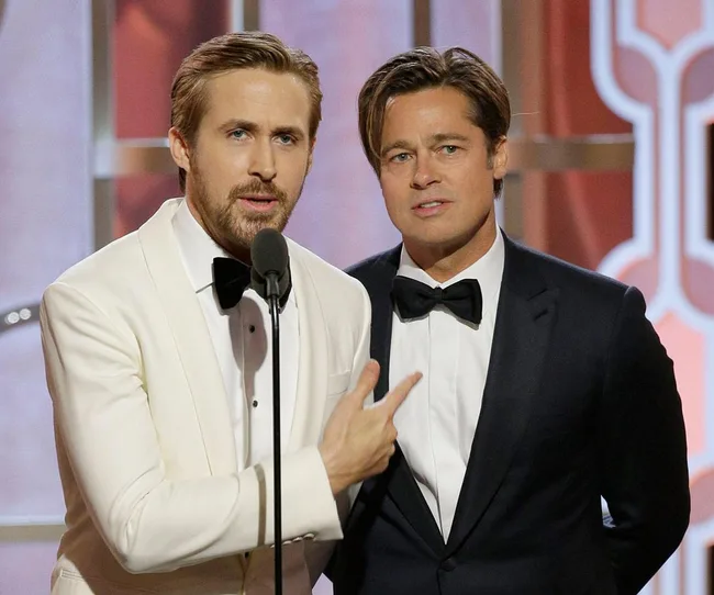 Two men in suits at an award ceremony, one in a white jacket speaking into a microphone.
