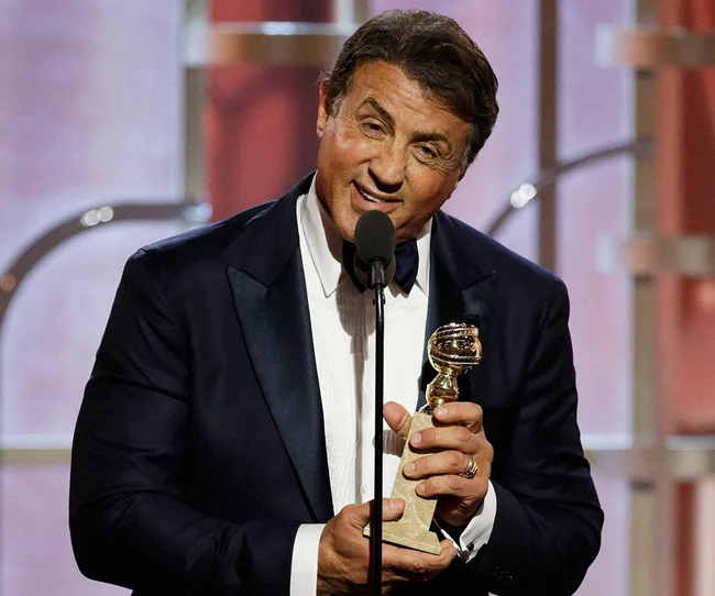 A man in a tuxedo holds a Golden Globe award and speaks into a microphone at an awards ceremony.