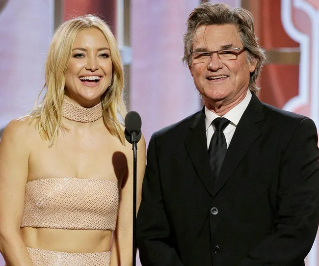 Two people smiling at an event, one wearing a sparkly dress, the other in a suit, standing by a microphone.