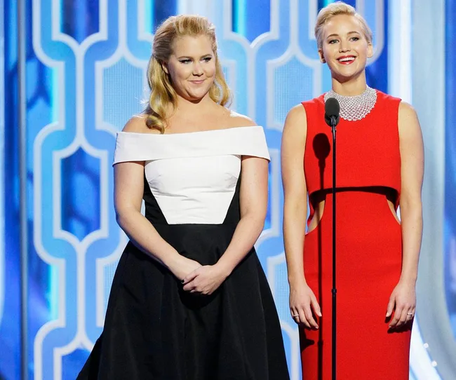 Two women in formal dresses stand on stage, smiling, with a microphone between them.