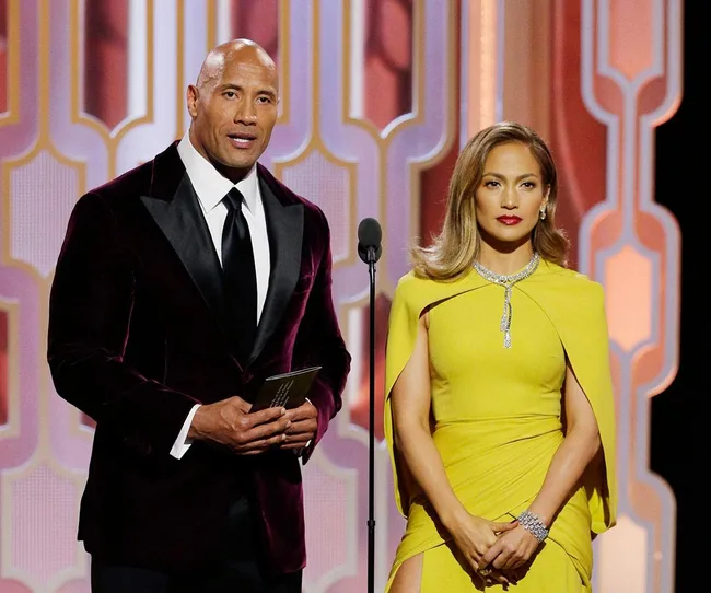 Two individuals on stage at an award event; one in a dark suit and another in a yellow gown.