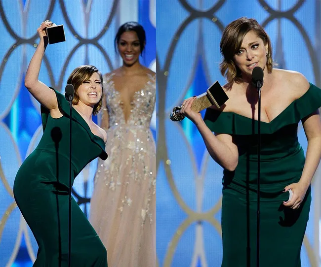 A woman in a green dress holds up an award on stage, smiling and speaking into a microphone.