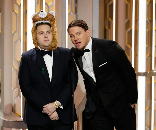 Two men in suits, one wearing a bear hat, speak at a microphone on an awards stage.