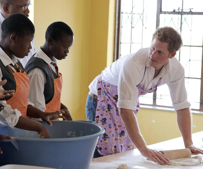 A person in an apron kneads dough on a table with students in school uniforms watching attentively.