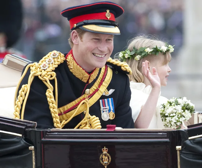 Royal in ceremonial uniform, smiling in a carriage with a girl wearing a flower crown and holding a bouquet.