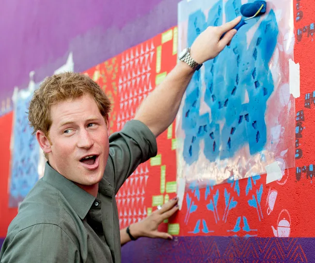 Man painting a colorful mural with a blue paint roller on a red and purple wall.