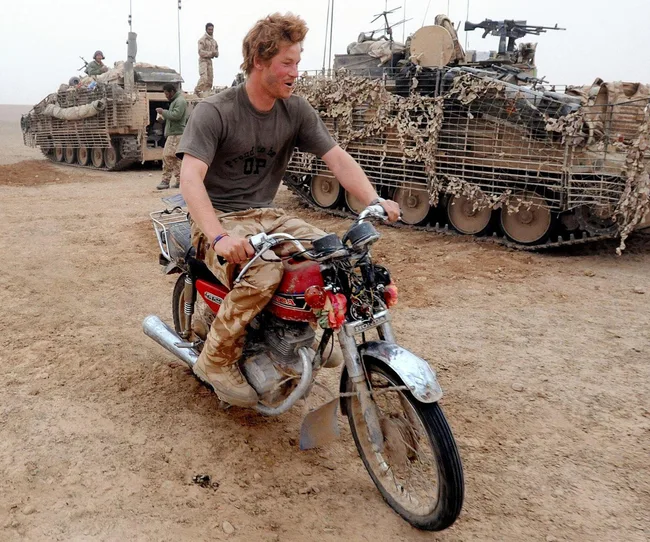 Man in military attire riding a motorcycle in a desert, with armored vehicles in the background.