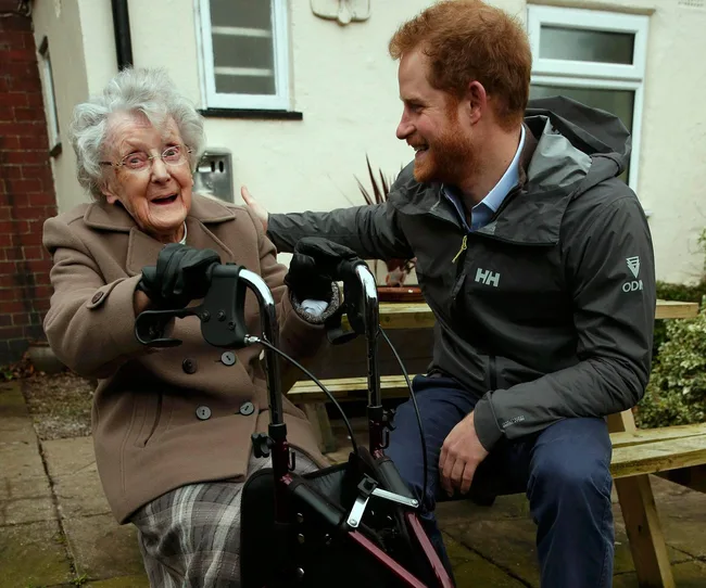 Senior woman with walker smiling at a man in a jacket, sitting outside on a bench.