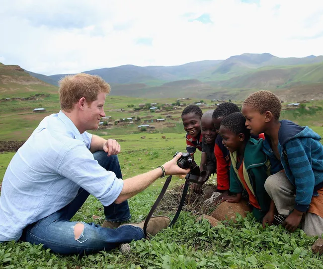 A man shows a camera to smiling children in a grassy field with hills in the background.