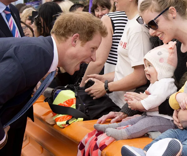 A man in a suit enthusiastically interacts with a smiling baby wearing a hat, surrounded by a crowd.