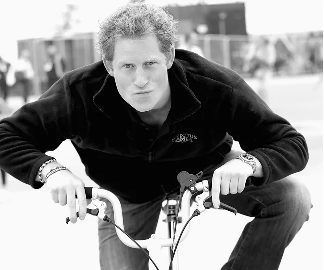 Man riding a bike, wearing a jacket with "Invictus Games" logo, looking towards the camera in a black and white photo.