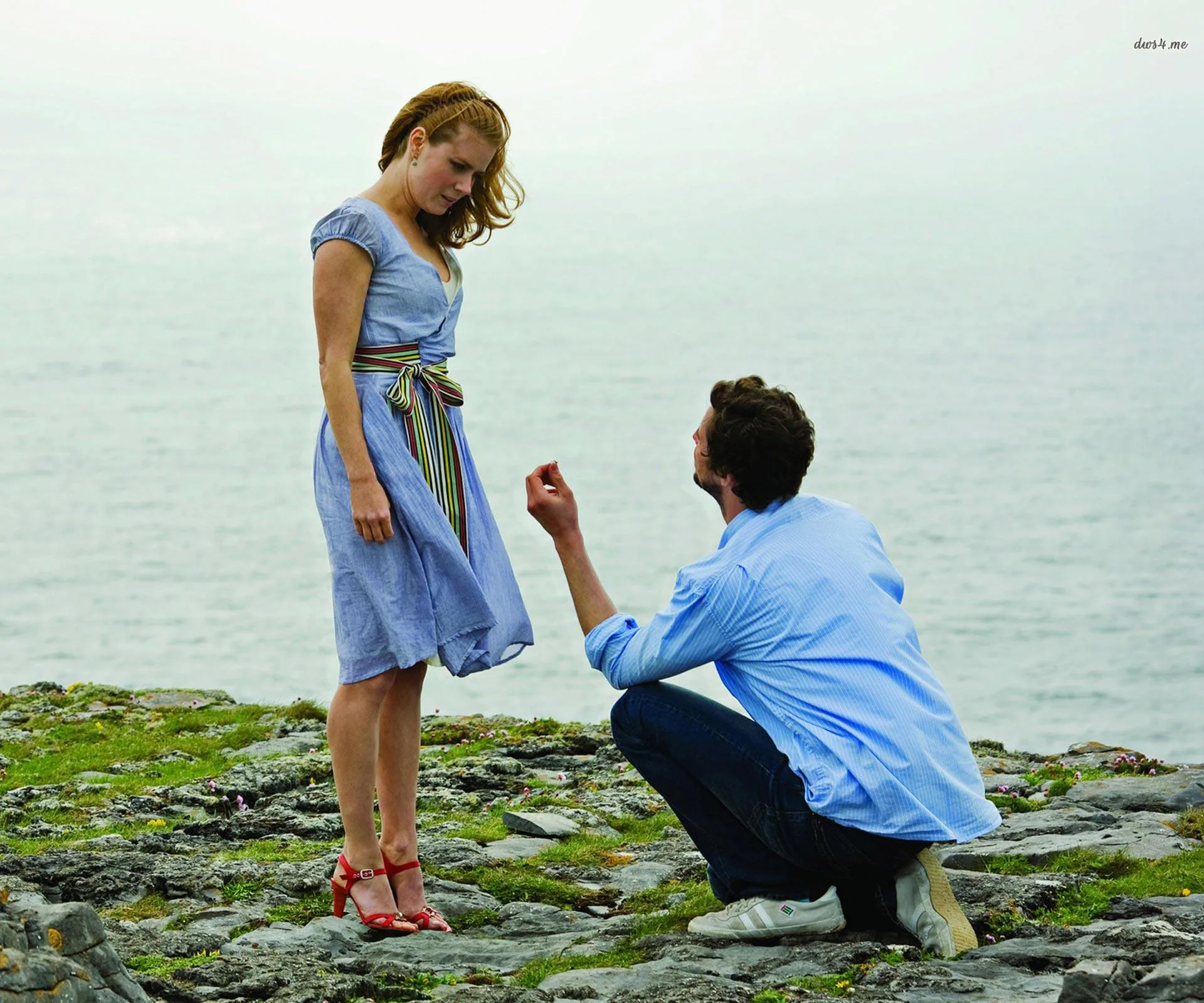 Man kneels with ring, proposing to woman on rocky shore with ocean in background.