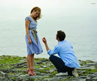 Man kneels with ring, proposing to woman on rocky shore with ocean in background.