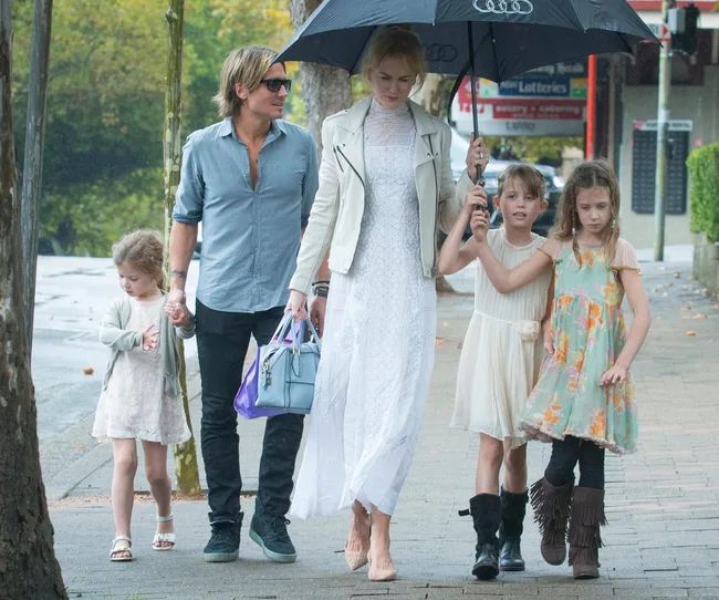 A family walking on a sidewalk; a woman holds an umbrella and a handbag, accompanied by a man and three children.