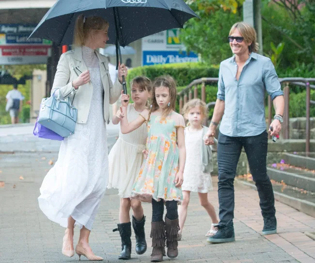 A man and woman walk with three girls under an umbrella on a rainy day in a casual setting.