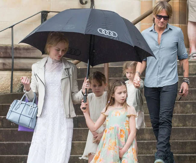 A family walks down steps under an umbrella; a woman in white holds a bag and a man in sunglasses holds a child's hand.
