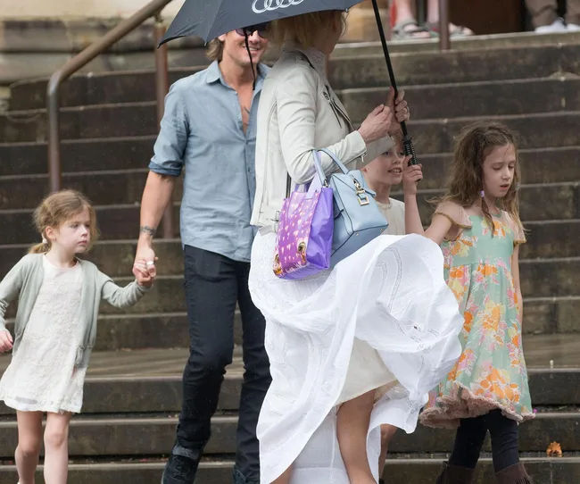 Family with children walking in rain; woman holds umbrella, man and young girls with floral dresses, windy atmosphere.