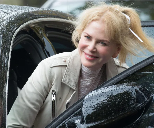 A woman with blonde hair in a ponytail, wearing a light jacket, smiles while stepping out of a car on a rainy day.