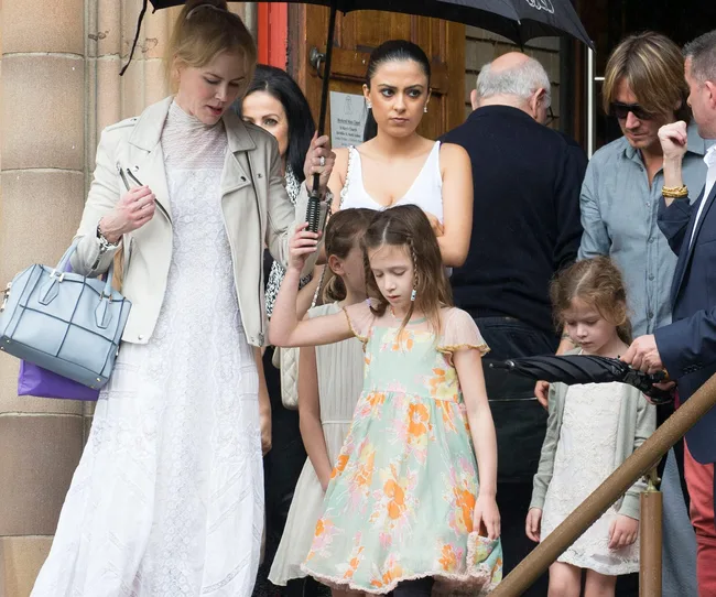 A woman in a white dress holds an umbrella with children and others around her, outside a building.