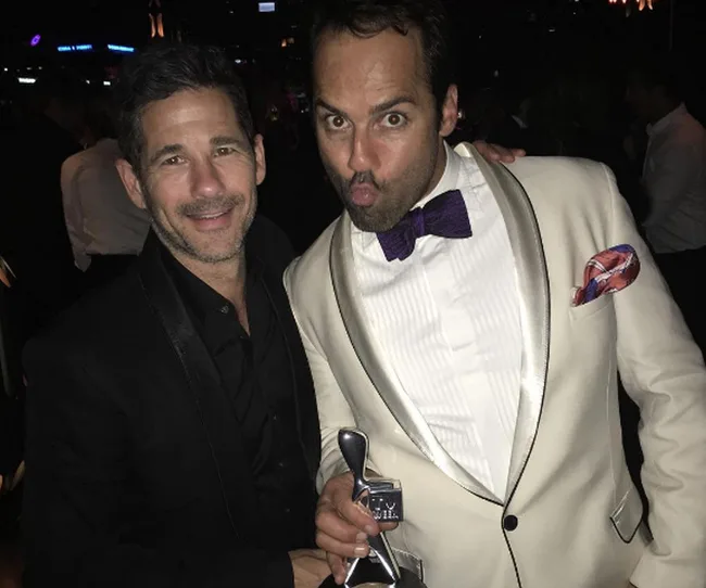 Two men in formal wear at a party; one wearing white suit holds a trophy, the other in black suit smiles at camera.
