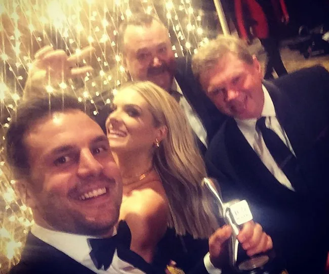 Selfie of four people in formal wear, smiling, with sparkling lights in the background. One holds a trophy.