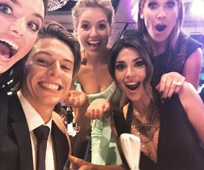 Group of five smiling people taking a selfie at an event, dressed in formal attire, looking excited and happy.