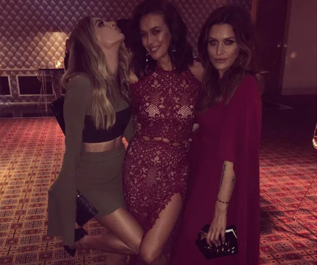 Three women in dresses posing for a group photo at an indoor event, with patterned walls and carpet.