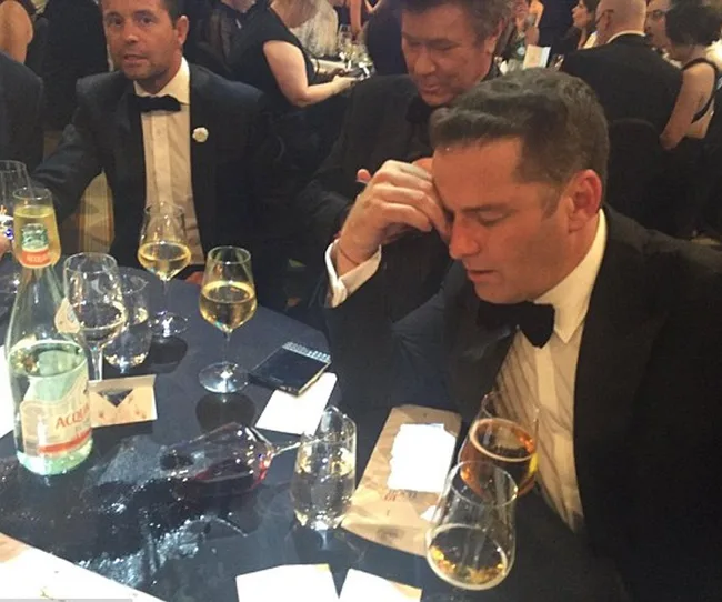 Three men in tuxedos sitting at a table with spilled drink and glasses at a formal event, one appearing tired or stressed.