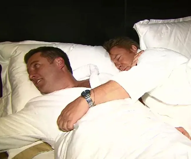 Two men in bathrobes lie in bed, one asleep hugging the other, who is awake and smiling.