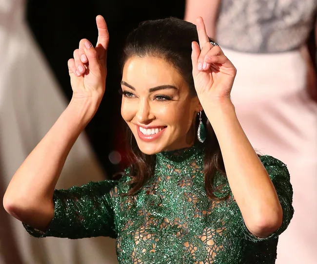 Smiling woman in a green lace dress making playful gesture with fingers raised, earrings visible, on red carpet.