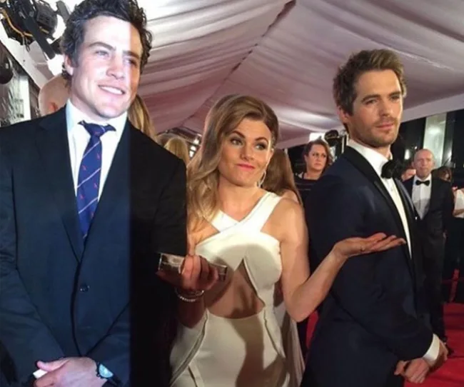 Three people in formal wear pose on a red carpet, with one woman in the middle making a shrugging gesture.