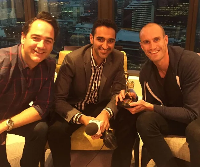 Three men smiling, seated indoors; one holds a microphone and another holds a trophy. Cityscape visible through window.