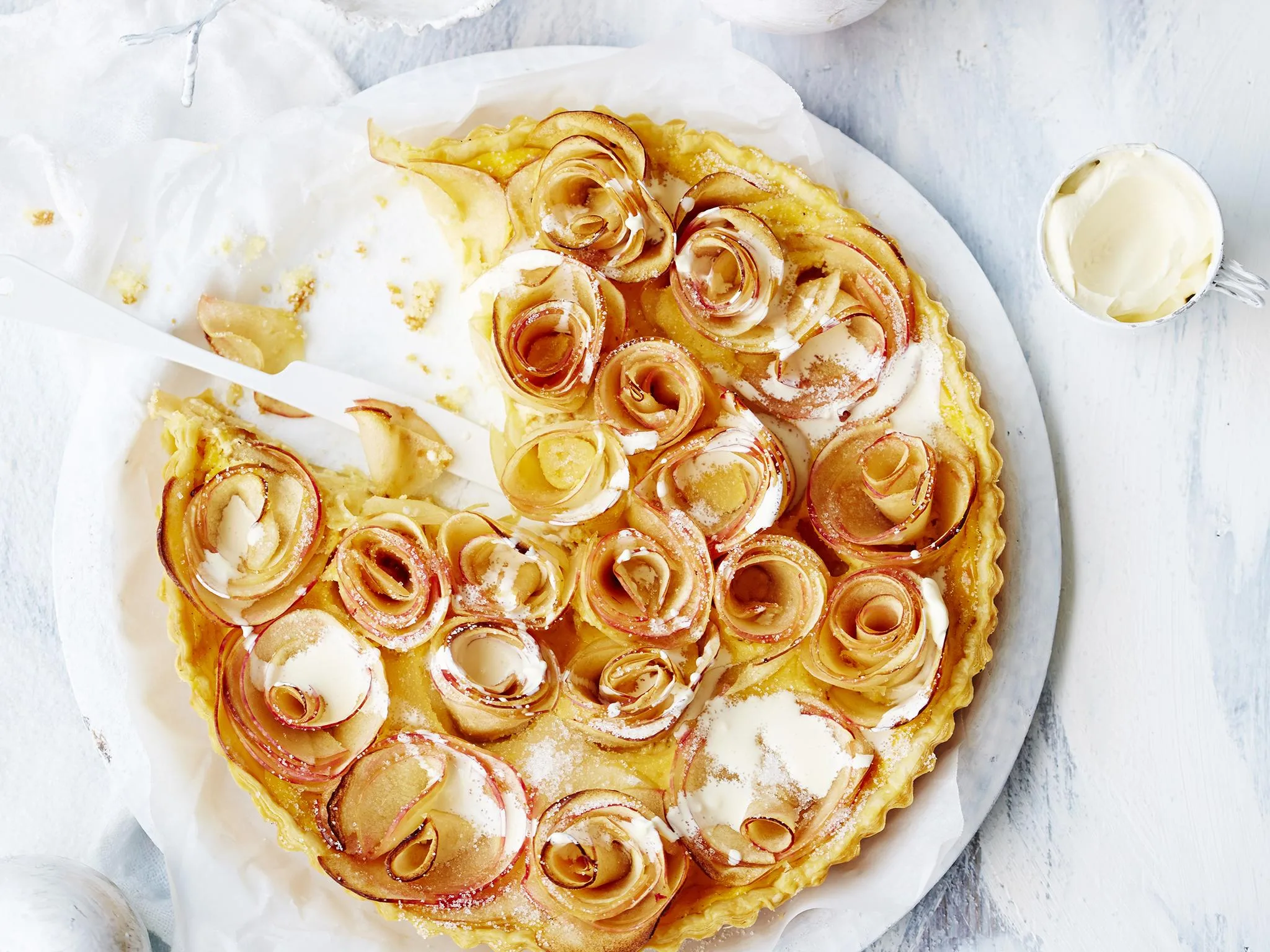 Apple tart with rose-shaped apple slices and cream on a white plate, partially sliced and ready to serve.