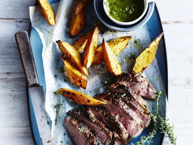 Grilled lamb with roasted potatoes and herbs on a blue platter with dipping sauce on the side.