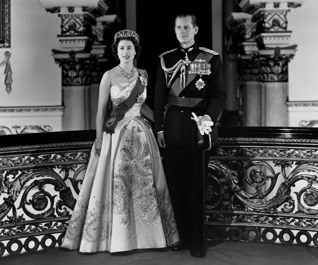 A royal couple in formal attire poses elegantly on an ornate staircase, with intricate metalwork in the background.