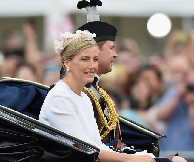 A woman in a white outfit with a floral hat sits beside a uniformed man in a carriage during a public event.