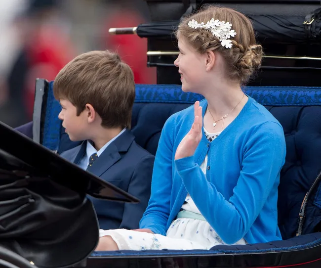 Two children sitting in a carriage, one wearing a blue suit, the other in a blue cardigan and floral hair accessory.