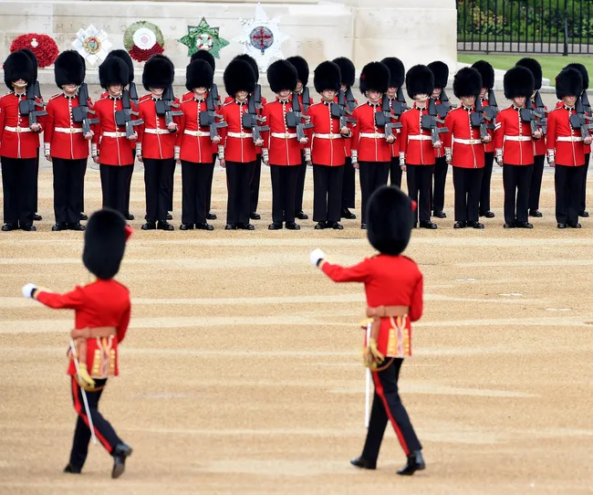 Ceremonial guards in red uniforms and bearskin hats stand lined up, with two guards marching in front.