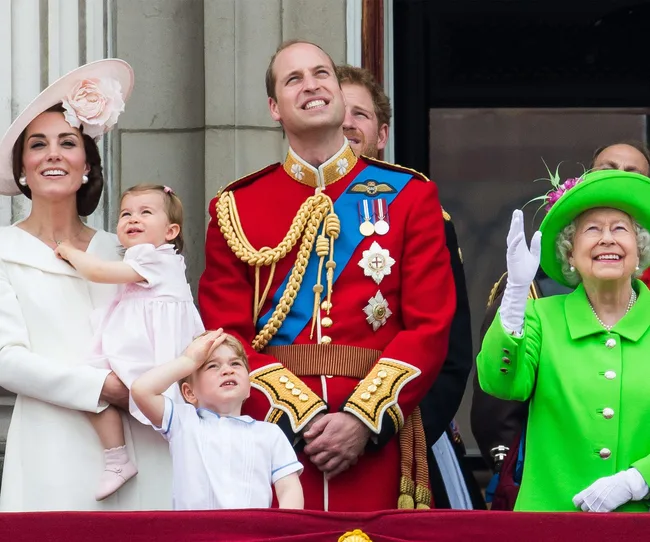 Royal family on a balcony during a public event, with a woman in white holding a child, a man in uniform, and an elderly woman in green.