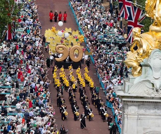 Aerial view of a parade celebrating a 90th birthday with large gold numbers, marching band, and crowd in London.